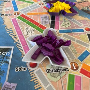 May include: A board game map of New York City featuring various coloured zones and landmarks like Times Square and Soho. Two star-shaped containers hold small plastic game pieces, one filled with yellow pieces and the other with purple pieces.