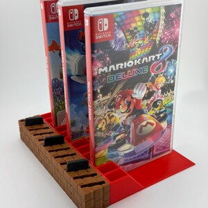 May include: A red and brown 3D-printed game cartridge holder with three Nintendo Switch game cases. The game cases are labelled "Mario Kart 8 Deluxe" and "Mario + Rabbids Sparks of Hope". The holder has a brick-like design.