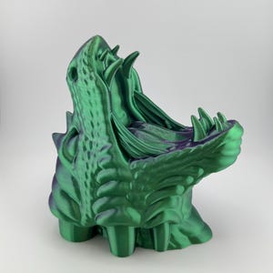 May include: A 3D-printed sculpture of a creature's open mouth, with sharp teeth and textured skin. The sculpture is green with purple accents, and features a detailed, stylized design. The open mouth reveals the interior of the sculpture.