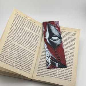 Pode incluir: Marcador de livros artesanal com uma ilustra&ccedil;&atilde;o em estilo de banda desenhada do rosto de uma personagem, em vermelho e preto. O marcador retangular est&aacute; colocado num livro aberto.