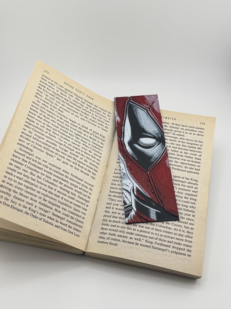 Pode incluir: Marcador de livros artesanal com uma ilustra&ccedil;&atilde;o em estilo de banda desenhada do rosto de uma personagem, em vermelho e preto. O marcador retangular est&aacute; colocado num livro aberto.