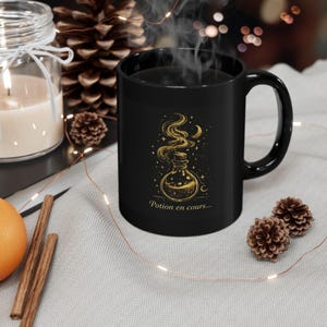 Peut inclure: Un mug noir en céramique avec une illustration dorée d'une fiole de potion, d'étoiles et d'un croissant de lune. Le mug porte l'inscription "Potion en cours..." et est entouré d'une bougie, de pommes de pin et de guirlandes lumineuses.