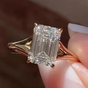 May include: A gold ring with a large, rectangular-cut diamond. The clear diamond is faceted, reflecting light. The ring has a split shank design. The brand name "VIVONA JEWEL" is engraved on the ring.