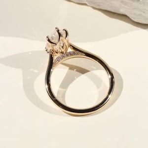 May include: A gold engagement ring with a large, clear gemstone and smaller accent stones. The ring has a delicate, detailed setting and a polished band. The ring is set against a neutral background, highlighting its design.