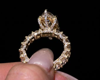 Può includere: Un anello color oro con un castone a forma di corona e numerose piccole gemme trasparenti. Anche la fascia dell'anello è adornata da gemme. L'anello è mostrato su uno sfondo nero.