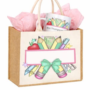 Op de afbeelding: Een bruine jute draagtas met een thema voor leraren. De tas heeft een leeg label, potloden, kleurpotloden en een mok met de tekst "Miss Thompson". De tas bevat roze vloeipapier en een bijpassende mok.
