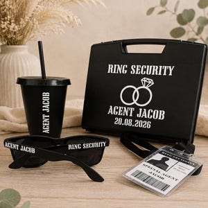 May include: A black plastic briefcase labeled RING SECURITY AGENT JACOB 20.08.2026 with a ring icon, a black tumbler with a straw labeled AGENT JACOB, black sunglasses labeled AGENT JACOB and RING SECURITY, and a security badge labeled SPECIAL AGENT JACOB.