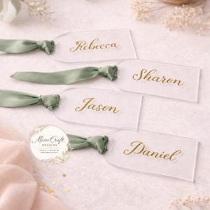 Puede incluir: Cuatro etiquetas rectangulares de acrílico transparente con la parte superior redondeada, cada una con una cinta de seda verde salvia atada. Los nombres Rebecca, Sharon, Jason y Daniel están impresos en cursiva dorada. Un logo dice Marce Craft.