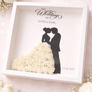Può includere: Una cornice bianca per matrimonio con la silhouette di una coppia e un abito fatto di rose bianche. Il testo "The Wedding of Katie & Karl" è sopra la coppia, con la data "13 Jul 2027" sotto.