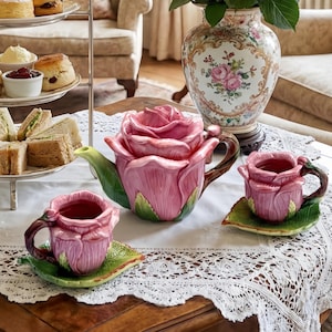 Peut inclure: Un ensemble de thé charmant avec une théière rose en forme de rose et des tasses assorties sur des soucoupes en forme de feuille verte. Un présentoir à trois niveaux présente des scones, des sandwichs et de la crème.