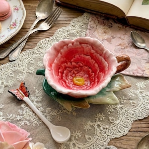 Puede incluir: Naturaleza muerta de temática floral con una taza de té en forma de flor roja y blanca, una cuchara de mariposa y un plato de macarons. La escena incluye cubiertos antiguos, un libro de botánica abierto y rosas rosas.