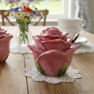 Puede incluir: Dos recipientes rosas de cerámica con forma de rosa y hojas verdes sobre una mesa de madera. Uno está abierto, revelando el interior. Un jarrón con flores y una taza de té están en el fondo.