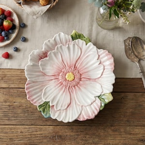 以下が含まれることがあります： ピンクと黄色のアクセントが付いた、大きな白いセラミック製の花型のサービングプラッター。プラッターは、ベリーのプレート、クロワッサン、花、銀食器に囲まれ、木製のテーブルの上に置かれています。