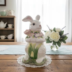 Peut inclure: Un pot à biscuits en céramique blanche en forme de lapin avec un motif floral. Le lapin tient une fleur rose et porte un collier floral. De l'herbe verte et des fleurs décorent la base. Le pot est posé sur un napperon en dentelle blanche.