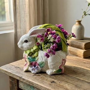 May include: A white ceramic bunny planter with a green handle and bow, filled with purple flowers. The bunny has detailed features and is decorated with floral accents. Perfect for Easter or spring decor.