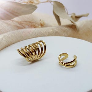 Könnte beinhalten: Ein Set aus zwei goldfarbenen Ohrklemmen. Eine Klemme hat ein breites, mehrschichtiges Design mit sieben parallelen, geschwungenen Bändern. Die zweite Klemme ist kleiner und hat ein einfaches, offenes Design mit zwei sich kreuzenden Bändern.