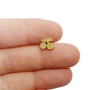 Peut inclure: Une petite breloque en métal doré en forme de paire de cerises. La breloque présente deux formes de fruits circulaires reliées par une courte tige sur le dessus, avec une finition de surface lisse et réfléchissante qui capte la lumière.