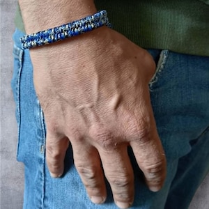 Op de afbeelding: Een geweven armband met een herhalend patroon van koningsblauwe, zwarte en metallic zilveren draden. Het accessoire heeft een getextureerd, gevlochten uiterlijk met een consistente dikte over de hele band, wat een gedetailleerd vlechtwerk toont.