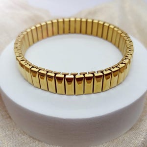Peut inclure: Un bracelet extensible doré composé d'une série de maillons métalliques rectangulaires et facettés reliés en une bande circulaire continue. La surface polie de chaque maillon reflète la lumière, créant un aspect métallique uniforme.