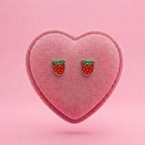 Puede incluir: Un par de pendientes pequeños tipo botón con forma de fresa, con cuerpos de esmalte rojo, puntos blancos diminutos y hojas verdes en la parte superior. Los pendientes se presentan sobre un cojín de terciopelo rosa en forma de corazón.