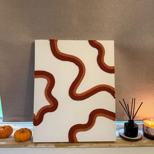 May include: Abstract canvas art featuring wavy lines in shades of brown and orange against a white background. The artwork has a minimalist design with textured lines. The piece is displayed on a shelf with pumpkins, a diffuser, and a candle.