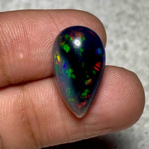 May include: A teardrop-shaped black opal gemstone with vibrant flashes of green, red, and yellow. The polished stone is held between fingers, showcasing its iridescent play-of-color. The opal's dark base enhances the brilliance of the internal colors.