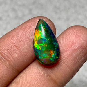 May include: A teardrop-shaped opal gemstone with a vibrant display of color. The stone features a dark base with flashes of green, orange, and blue. The polished surface reflects light, showcasing the opal's internal play-of-color.
