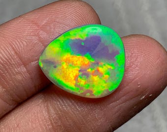 May include: A vibrant, polished opal gemstone with a mix of colors. The stone displays a play of color with shades of green, yellow, and purple. The opal is held in a hand, showcasing its unique, natural beauty. The gemstone is a unique piece of jewelry.