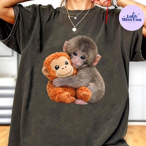 May include: Dark olive green t-shirt featuring a graphic of a baby monkey hugging an orange stuffed monkey. The image is centered on the shirt. The text "Lola Miss You" is in a purple circle in the upper right corner.
