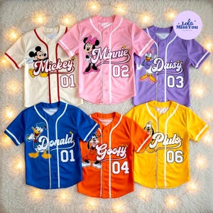 May include: Six colorful baseball jerseys featuring Disney characters. The jerseys are cream, pink, purple, blue, orange, and yellow. Each jersey has a character's name and number on the front, including Mickey, Minnie, Daisy, Donald, Goofy, and Pluto. The jerseys have white trim.