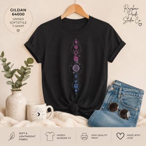 Puede incluir: Una camiseta negra de manga corta con un gráfico vertical de planetas y cuerpos celestes en tonos rosa, morado y azul. La camiseta tiene cuello redondo y se muestra en una percha de madera. El texto en la camiseta incluye GILDAN 64000.