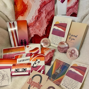 Op de afbeelding: Een verzameling lesbische pride-artikelen, waaronder stickers, buttons, kunstprints en een cassettebandje. De items tonen de kleuren van de lesbische vlag, met tekst zoals "Lesbian" en "Sapphic".