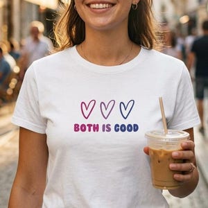 Könnte beinhalten: Weißes T-Shirt mit dem Text „BOTH IS GOOD“ und drei Herzsymbolen in Rosa, Lila und Blau. Die Person trägt das T-Shirt und hält ein Getränk in einem transparenten Plastikbecher mit Strohhalm.