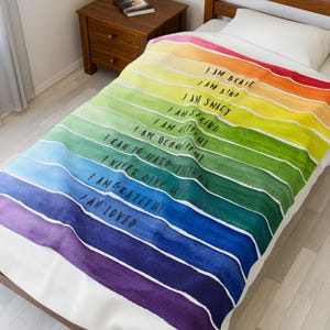 Könnte beinhalten: Eine farbenfrohe Decke mit einem Aquarell-Regenbogen-Design. Die Decke hat horizontale Streifen in verschiedenen Farben, darunter Rot, Orange, Gelb, Grün, Blau und Lila. Der Text in schwarzer Schrift lautet: "I AM BRAVE, I AM KIND, I AM SMART, I AM STRONG, I AM HELPFUL, I CAN DO HARD THINGS, I NEVER GIVE UP, I AM GRATEFUL, I AM LOVED."