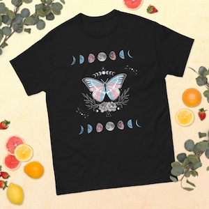 Könnte beinhalten: Schwarzes T-Shirt mit einem Schmetterlingsdesign in den Farben der Transgender-Pride-Flagge, umgeben von Mondphasen und floralen Elementen. Ein grafisches T-Shirt mit einem himmlischen Thema.