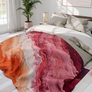 Könnte beinhalten: Eine farbenfrohe Decke mit einem abstrakten Design in Orange-, Weiß- und verschiedenen Rosa- und Rottönen. Die Decke ist auf einem Bett mit neutralfarbener Bettwäsche drapiert. Das Design hat ein strukturiertes, fließendes Aussehen.