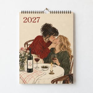May include: A 2027 wall calendar featuring an illustration of two people kissing at a round table. The scene includes a bottle of Vin Rouge, two glasses of red wine, a small bowl of olives, a lit candle, and a vase of flowers on a checkered cloth.