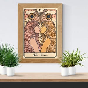 May include: A framed art print of a tarot card, "The Lovers," featuring two women kissing. The artwork has a vintage style with warm colors, sunflowers, and the number 11 at the top. The frame is made of wood.