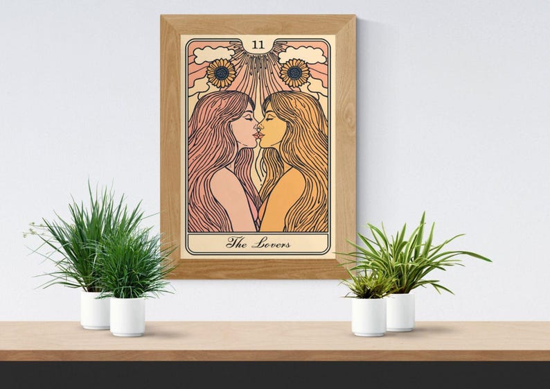 May include: A framed art print of a tarot card, "The Lovers," featuring two women kissing. The artwork has a vintage style with warm colors, sunflowers, and the number 11 at the top. The frame is made of wood.