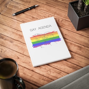 May include: A white notebook with the words "GAY AGENDA" printed on the cover. The notebook features a rainbow-colored design with a line drawing of two people kissing. A black pen and a mug are also visible.