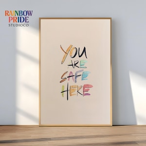 May include: A framed art print featuring the phrase "YOU ARE SAFE HERE" in multi-coloured, handwritten lettering. The artwork has a beige background and a gold-coloured frame. The upper left corner has the text "RAINBOW PRIDE STUDIOCO".