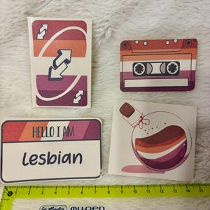 Könnte beinhalten: Vier Aufkleber mit Designs der lesbischen Pride-Flagge. Einer ist eine Uno-Reverse-Karte, ein weiterer eine Kassette, ein Namensschild mit "Hello I am Lesbian" und eine Zaubertrankflasche.