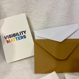 Könnte beinhalten: Eine weiße Karte mit dem Text "VISIBILITY MATTERS" in Regenbogenfarben, neben einem braunen und weißen Umschlag. Die Karte und die Umschläge liegen auf einer weißen, strukturierten Oberfläche.