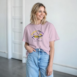Può includere: T-shirt rosa chiaro con la scritta "Vibes Only" e una grafica di geco nero, giallo e viola. Il testo "Too Far to Camouflage" è sotto. La maglietta è abbinata a jeans blu.
