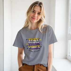 Könnte beinhalten: Ein graues T-Shirt mit dem Text "THEY THEM VIBES" in einer farbenfrohen Retro-Schriftart. Das Shirt enthält auch den Satz "BEYOND THE BINARY" in einer kleineren Schriftart. Das Design hat gelbe, lila und weiße Farben.