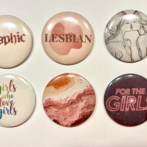 Puede incluir: Un conjunto de seis botones redondos. Los botones presentan varios diseños y textos, incluyendo "Sapphic", "Lesbian", un boceto de dos mujeres besándose, "girls who love girls", un patrón abstracto y "FOR THE GIRLS".
