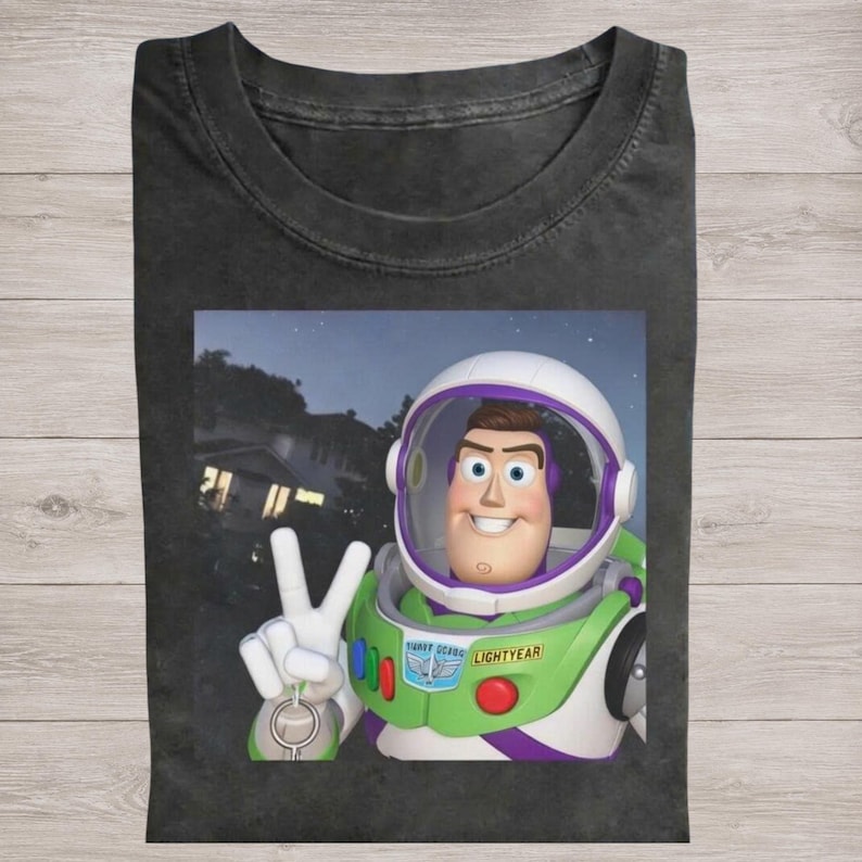 May include: Dark gray t-shirt featuring a graphic of Buzz Lightyear from Toy Story. Buzz is wearing his space suit and giving a peace sign. The shirt is a crew neck.