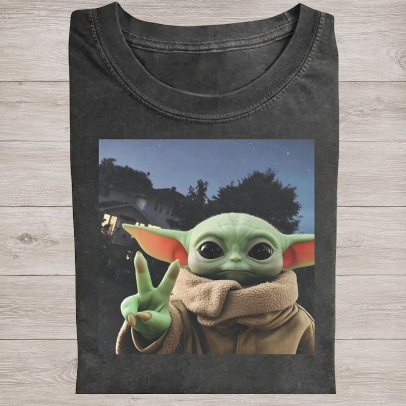 Pode incluir: Camiseta cinza escuro com um gr&aacute;fico de um personagem alien&iacute;gena verde fazendo o sinal de paz. O alien&iacute;gena est&aacute; usando um casaco marrom e um cachecol bege. O fundo mostra um c&eacute;u noturno com uma casa e &aacute;rvores.