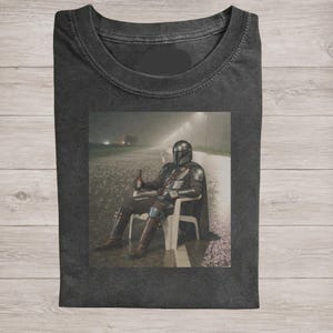 May include: A charcoal gray crew neck t-shirt featuring a graphic print of a armored character in a helmet sitting on a white plastic chair on a rainy road at night, holding a brown bottle in one hand while wearing metallic armor plating.