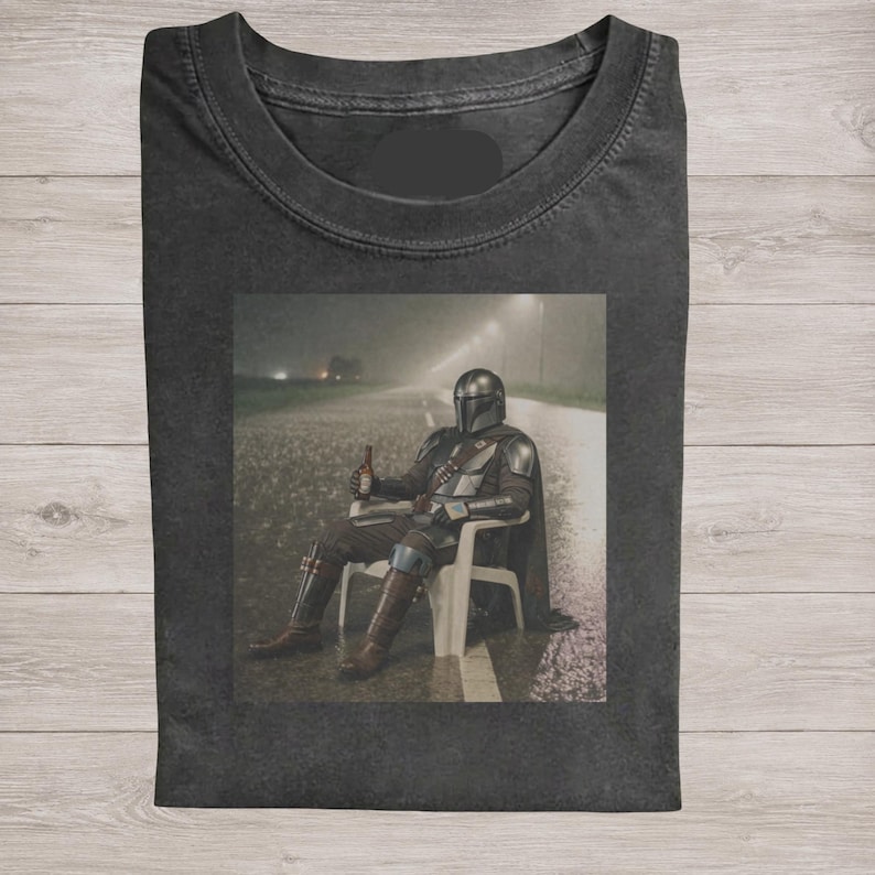 May include: A charcoal gray crew neck t-shirt featuring a graphic print of a armored character in a helmet sitting on a white plastic chair on a rainy road at night, holding a brown bottle in one hand while wearing metallic armor plating.